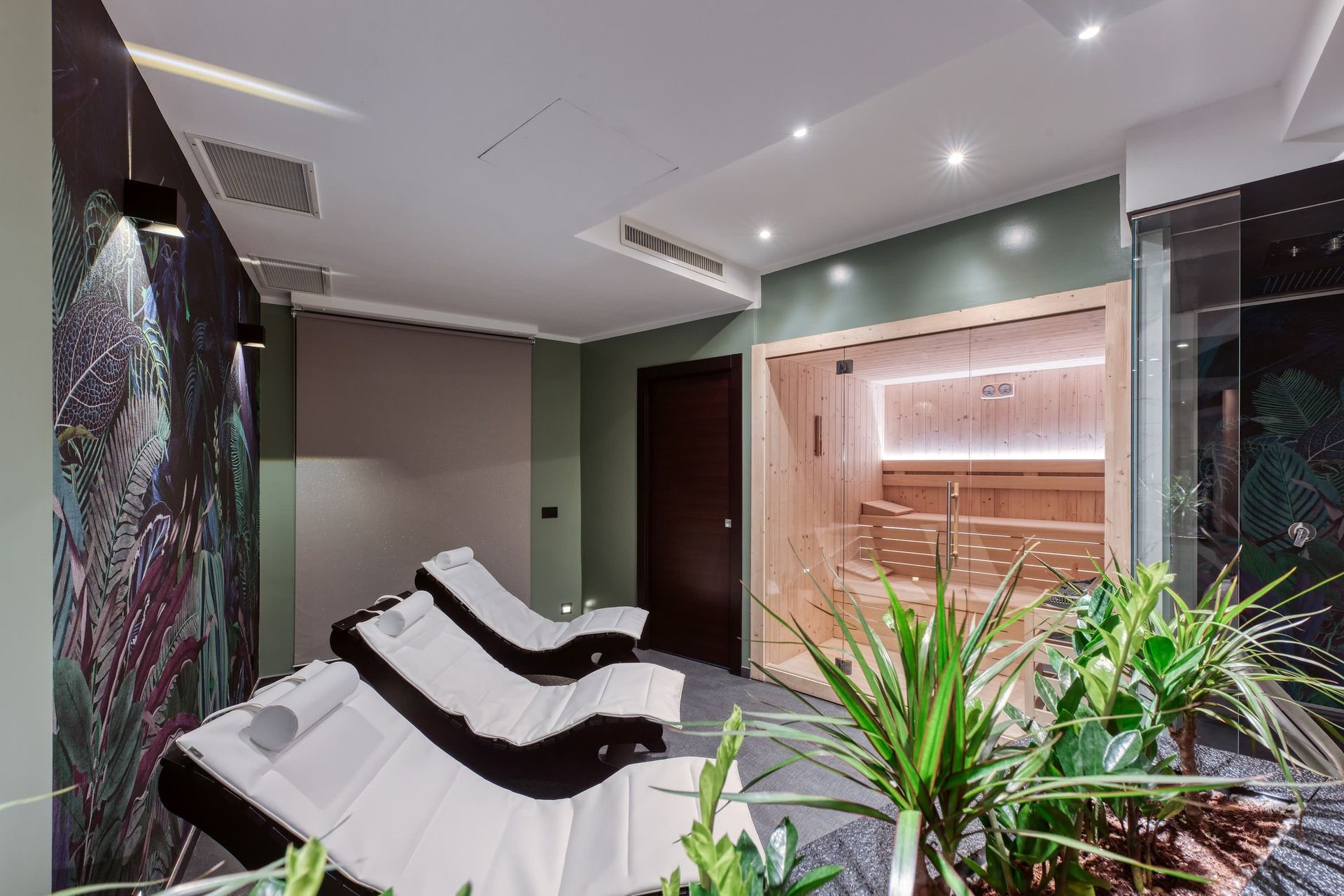 La zona relax con le chaise longue, la parete tropicale e la sauna dietro il vetro